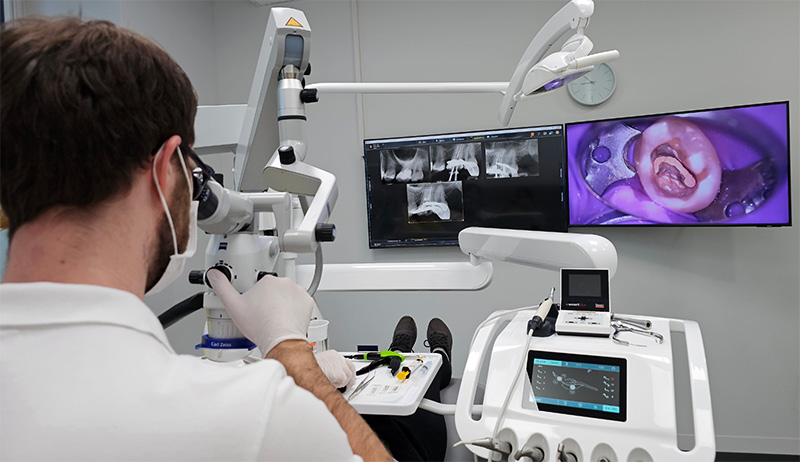Zahnarzt arbeitet mit einem Dentalmikroskop, w&auml;hrend auf Monitoren vergr&ouml;&szlig;erte Aufnahmen eines Zahns und R&ouml;ntgenbilder zu sehen sind