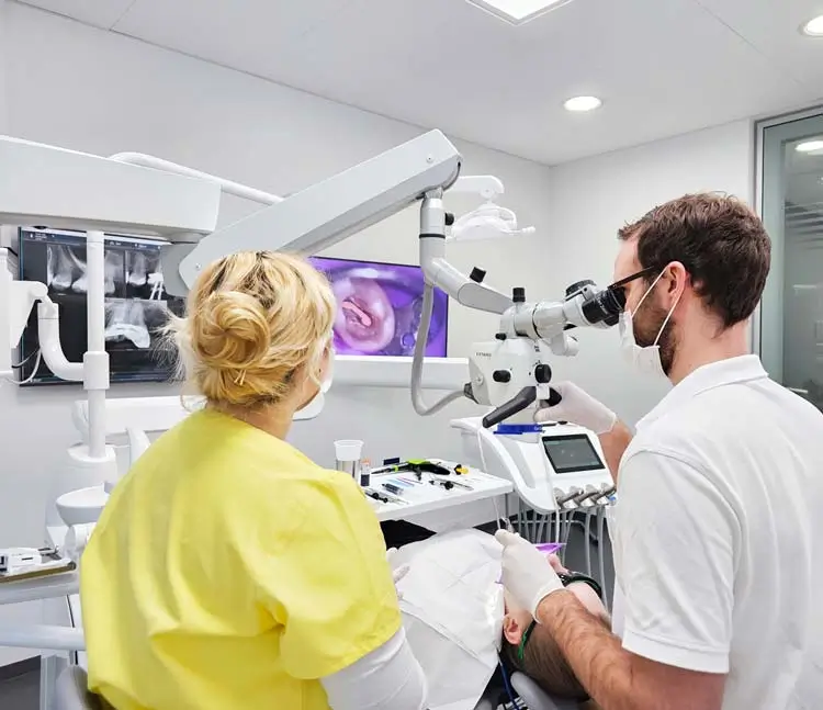Zahnarzt mit Assistenz arbeiten an einem Patienten. Einer benutzt ein Mikroskop, während die andere danebensteht. Im Hintergrund sind Röntgenbilder und ein Monitor zu sehen.